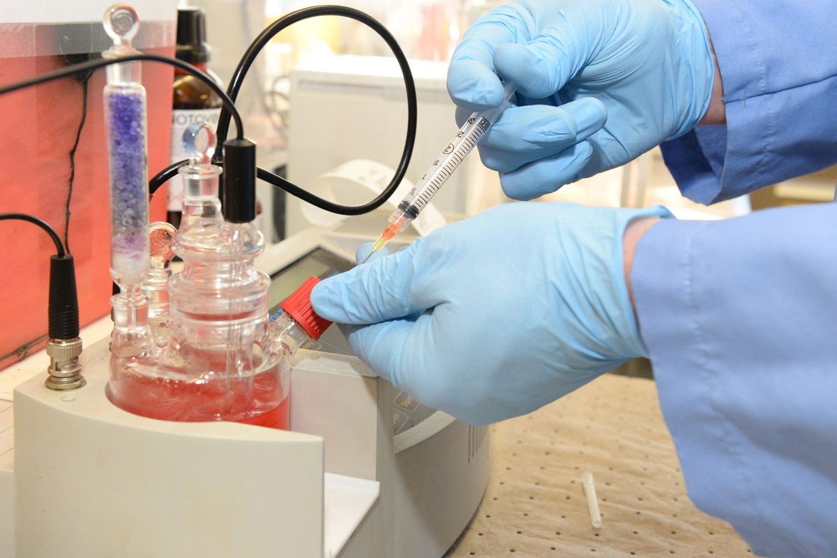 Gloved hands push a syringe needle through the port of a glass Karl Fischer titration cell holding pink reagent, beside a drying tube filled with blue granules.