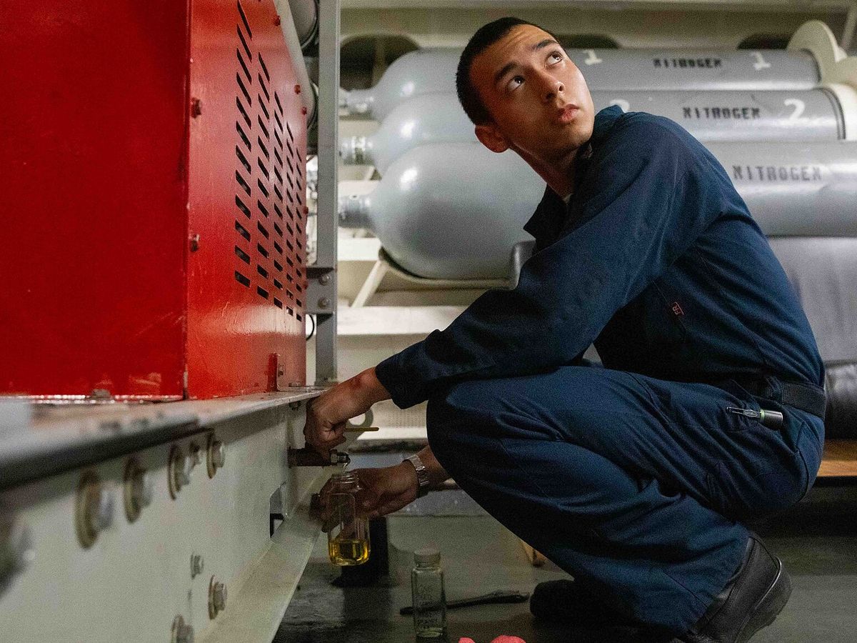 A sailor in navy coveralls crouches beside a red generator enclosure, holding a small glass jar of amber oil under a valve low on the machine base. A second, empty jar stands on the deck.