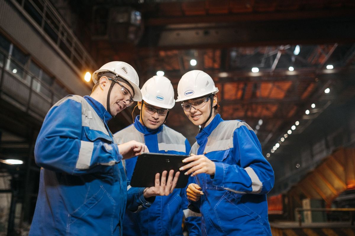 Three engineers in white hard hats and blue workwear look at a tablet together on the floor of a large industrial plant.
